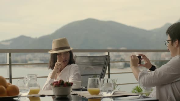 Young Woman is Taking Photo of Young Woman Friend Sitting at Table Outdoors While Traveling Rbbro