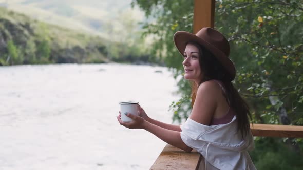 Portrait of a Beautiful Brunette with a Cup of Tea on a Background of Mountains and a River