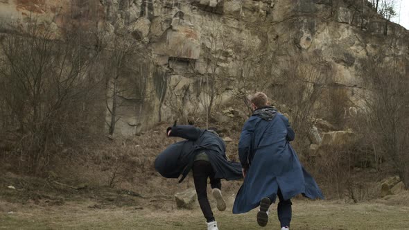 Two Young Guys Running Forward. Guys In Raincoats On Rock Background. View From The Back.