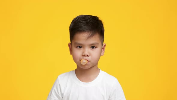 Little Asian Boy Chewing And Blowing Bubblegum Over Yellow Background