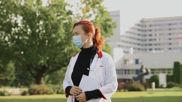 Female Doctor in Surgical Face Mask Holds Papers and Looks Around