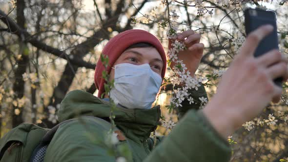 Man in a Medical Mask Takes a Selfie on the Phone with a Blossoming Apple Tree at Sunset