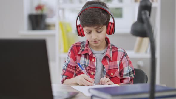 Positive Middle Eastern Schoolboy Doing Homework Listening Music in Headphones