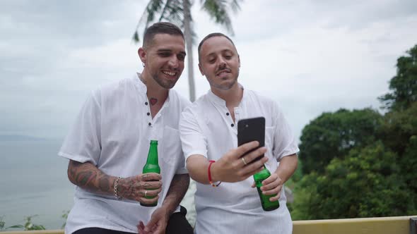 Two Happy Men Gay Couple Honeymooners Holding Beer and Watching Phone Pictures During Summer