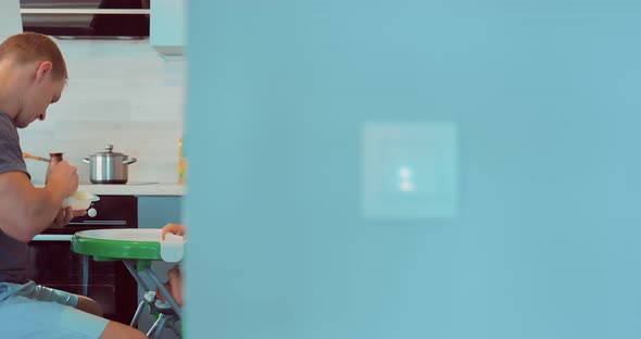 Handsome Young Father Feeding to His Baby Food in the Kitchen