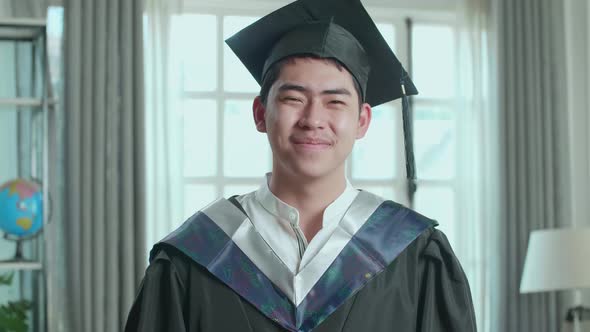 Asian Man Wearing A Graduation Gown And Cap Smiling To Camera In Living Room