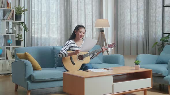 Smiling Asian Woman Composer Looking At Paper In Hand And Shaking Her Head Before Playing A Guitar