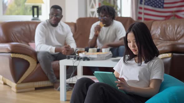 Confident Young Millennial Asian Woman Messaging on Tablet As Relaxed African American Men Chatting