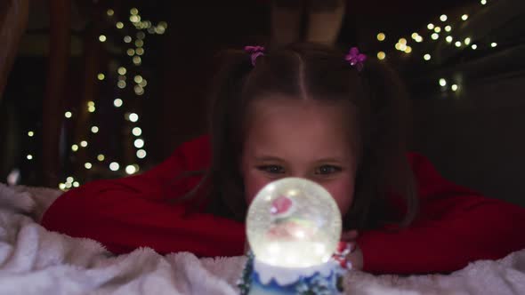 Caucasian girl smiling and looking at snow globe while lying under blanket fort during christmas