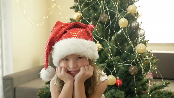 Portrait of a Sad Little Girl in a Santa Hat Against the Background of a Christmas Tree.