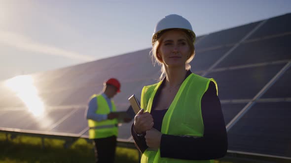 Portrait of a Female Solar Engineer Standing Looking at the Camera Another Man Near the Panels and