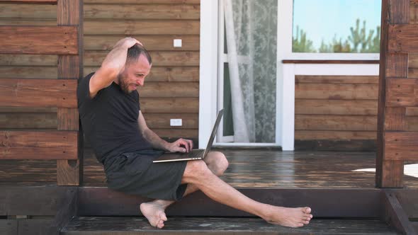 A Man in Shorts is Working on a Laptop Sitting on the Terrace Near the House A Businessman in a