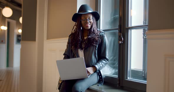 Portrait of Stylish African Woman Sitting on Windowsill Working on Laptop