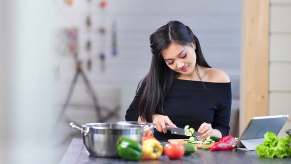 Medium Shot Pretty Smiling Young Asian Woman Cutting Cucumber Sitting at Table in Kitchen