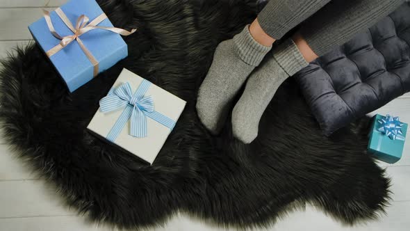 Female Legs in Warm Socks Rest Comfortably on a Gray Fur Skin Next To Blue and White Gift Boxes with