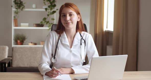 Young Pensive Woman Doctor Wears Medical Uniform, Sits in Clinic in Office with Laptop, Looks Out