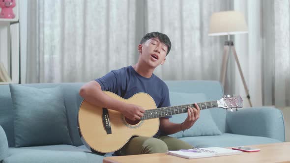 Asian Boy Composer With Notebook On Table Singing And Playing Guitar At Home