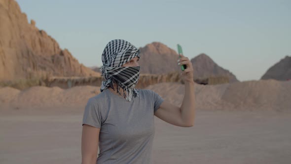 Woman in Checkered Keffiyeh Trying to Find Mobile Network Using Mobile Phone Standing in Desert