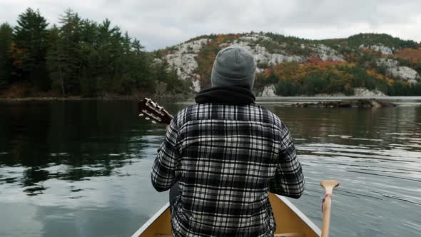 Man plays Ukelele in Canoe in Autumn Leaf Color Forest in slow motion on Canada lake. Wide Pan of Fa