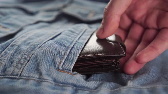 hand pulls a brown wallet out of the pocket of a blue denim jacket.
