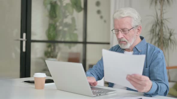 Senior Old Man Reading Documents While Using Laptop in Office