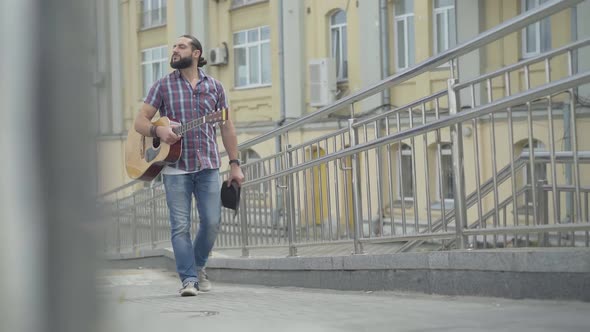 Thoughtful Caucasian Man Walking Along European City Street with Guitar and Hat. Portrait of Sad