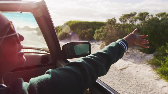 Mixed race woman driving on sunny day in convertible car waving arm and smiling