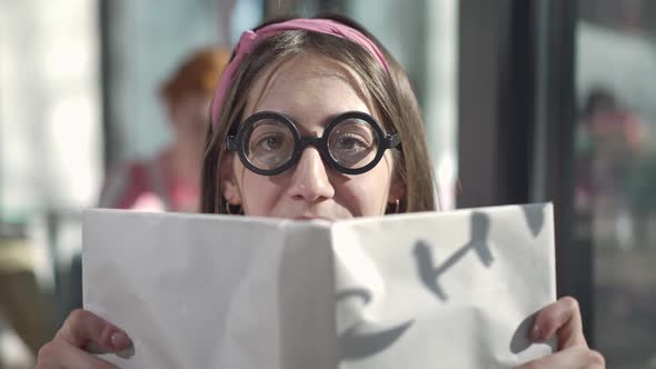 Closeup of Cheerful Nerd Young Caucasian Woman in Eyeglasses Showing Over Book and Hiding