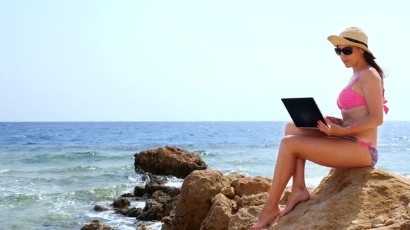 Young Woman in Bright Pink Swimsuit and Sunglasses, Uses Laptop, Sitting on Stone on Beach By the