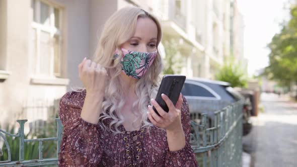 A Middleaged Caucasian Woman in a Face Mask Works on a Smartphone and Celebrates in an Urban Area