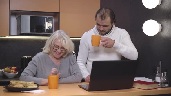 Senior Caucasian Husband and Wife Surfing Internet on Laptop in Kitchen and Talking. Portrait of