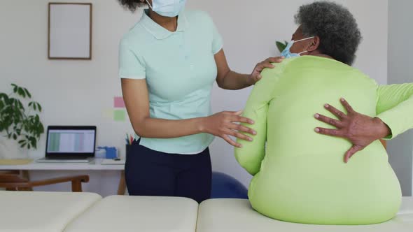 African american female physiotherapist wearing face mask helping senior female patient exercise