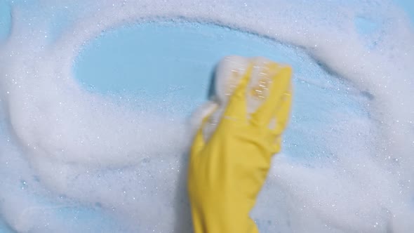 Hand in a Yellow Rubber Glove Holds a Cleaning Sponge and Wipes a Soapy Foam on a Blue Background