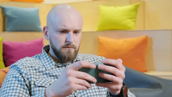 Close Up of an Young Man Sits on a Bean Bag While Playing Online Using His Phone