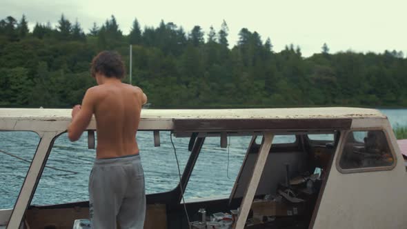 Topless young male walks into frame scrapes old mastic from timber boat cabin roof planking.