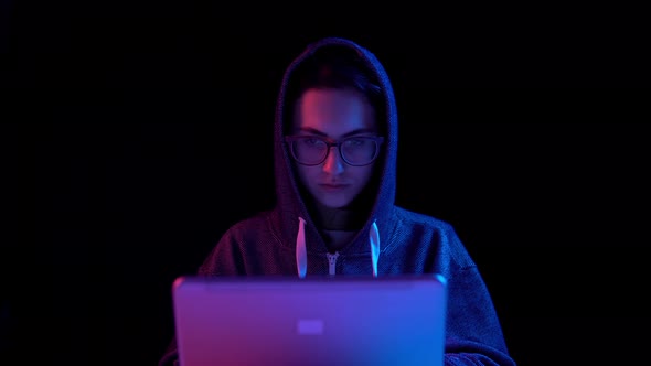 Young Woman in a Hood with a Laptop. Hacker Makes a Hack Through a Laptop. Blue and Red Light Falls
