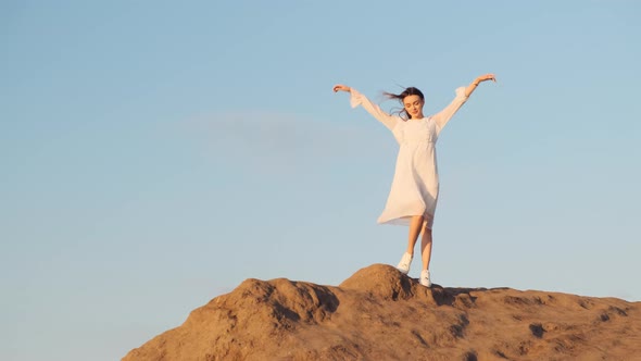 Slender Sexy Model Woman in White Long Dress and White Sneakers Stands on Top of Mountain and Raises