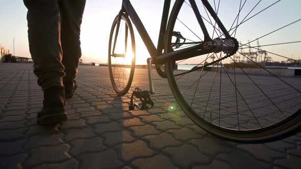 Young Hipster Man Walking with Bicycle During Sunset or Sunrise with Sea Port on Background
