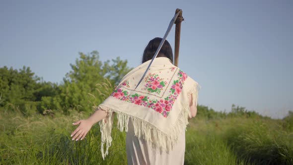 Back View of Overweight Woman in Traditional Shawl Walking on Summer Field with the Scythe