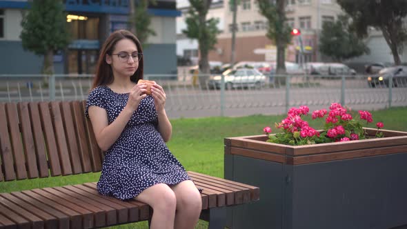 A Young Pregnant Girl is Eating a Burger While Sitting on a Bench in the Park