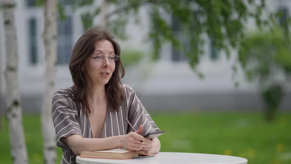 Female Teacher is Working with Book and Mobile Phone in Open Terrace of Cafe