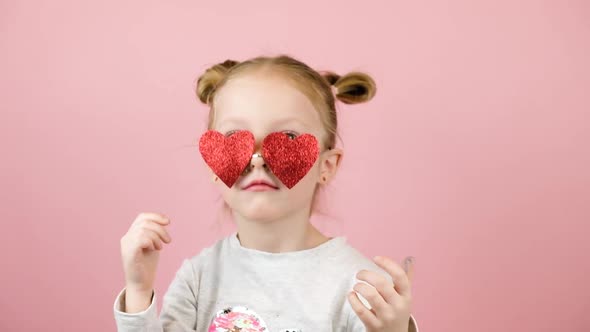 Funny Little Blonde Girl Smiling and Playing with Red Heart Shape Sunglasses on Pink Background
