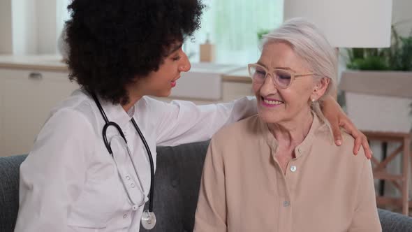 Afro American Woman Doctor Supports Senior Woman with Hand on Shoulder While Sitting on Sofa