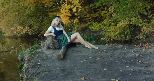 Happy Blonde in Slavic Costume Admires Autumn Nature Near River at Tree Stem