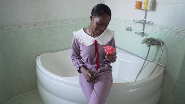 Portrait of Charming Romantic African American Woman in Pajamas Sitting on Bathtub with Red Rose