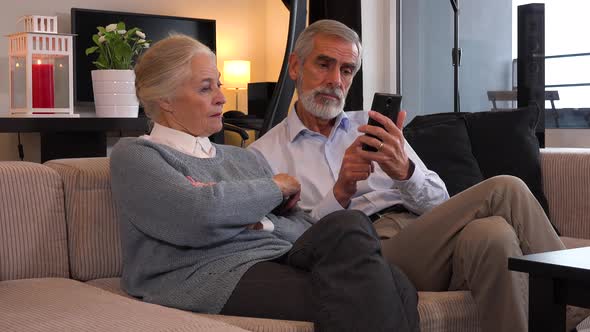 An Elderly Couple Sits on A Couch in An Apartment and Uses Phone Together and Talks