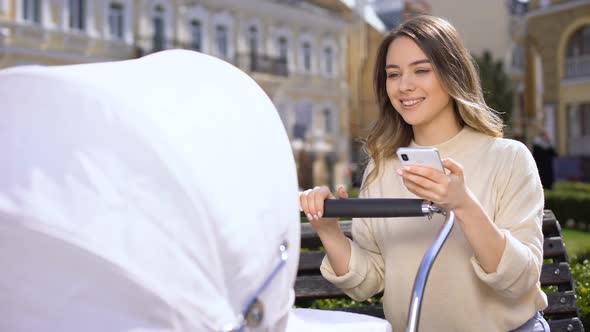 Joyful Young Mother Reading Internet Forum on Smartphone Swinging Baby Carriage