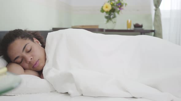 Attractive African American Woman Sleeping Under White Blanket in the Light Room on the Background