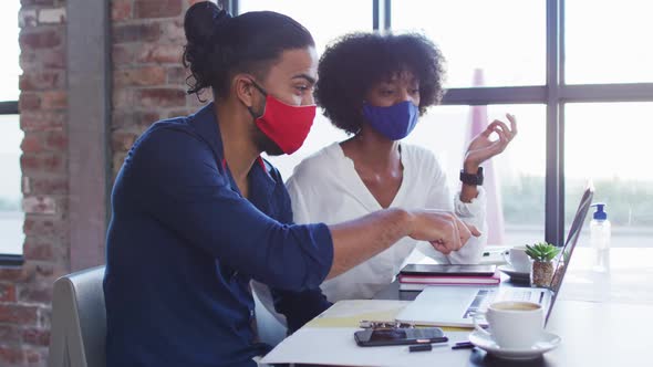 Diverse couple wearing face masks sitting in cafe using laptops and talking