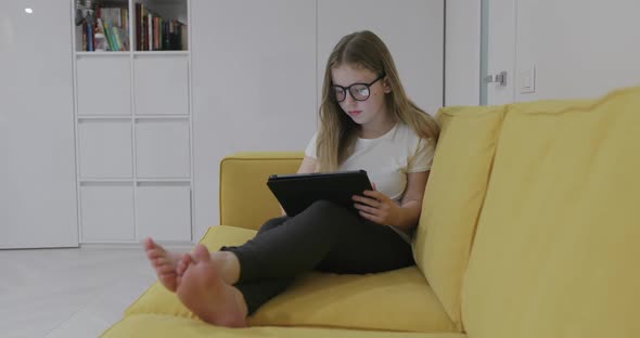 Barefoot Girl in Glasses Sitting on Sofa and Watching Video on Tablet While Resting at Home During